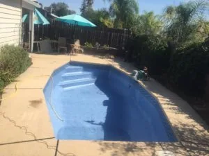 Empty in-ground swimming pool with blue interior, surrounded by tan concrete deck, black wooden fence, turquoise umbrellas, and palm trees.