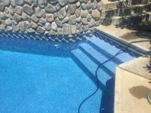 Swimming pool with blue water and stone wall, concrete deck with steps and equipment hose