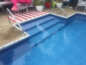 Blue swimming pool with built-in steps and metal railings, pink and white striped lounge chair visible poolside with plants and patio furniture