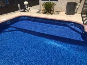 Backyard swimming pool with bright blue interior lining and red brick coping, surrounded by concrete patio and desert landscaping