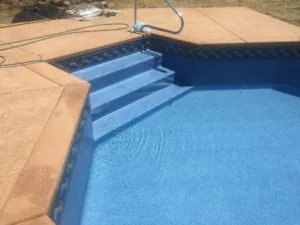 Residential swimming pool with blue plaster interior and light-colored stone deck, featuring interior steps and a hose on the deck edge.