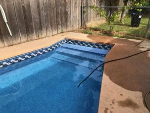 In-ground swimming pool with blue water and stepped interior, surrounded by concrete deck and weathered wooden fence