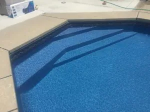 Indoor swimming pool with blue textured floor and dark water lane markings, beige deck surround with product box visible