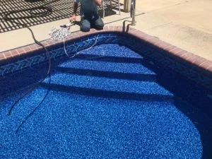 Person kneeling beside swimming pool with blue interior surface and decorative tile border, holding hose with coiled rope on concrete deck