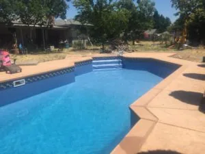 Residential swimming pool with bright blue water and tan concrete deck surrounded by rural property with trees and outbuildings