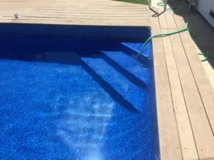 Swimming pool with blue interior and stairs visible, wooden deck surrounding the pool edge with green hose