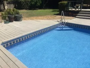 In-ground swimming pool with blue water and decorative border, wooden deck with potted plants, fenced backyard and metal structure visible