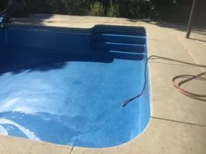 Empty swimming pool with blue interior and built-in steps, concrete deck, and garden background