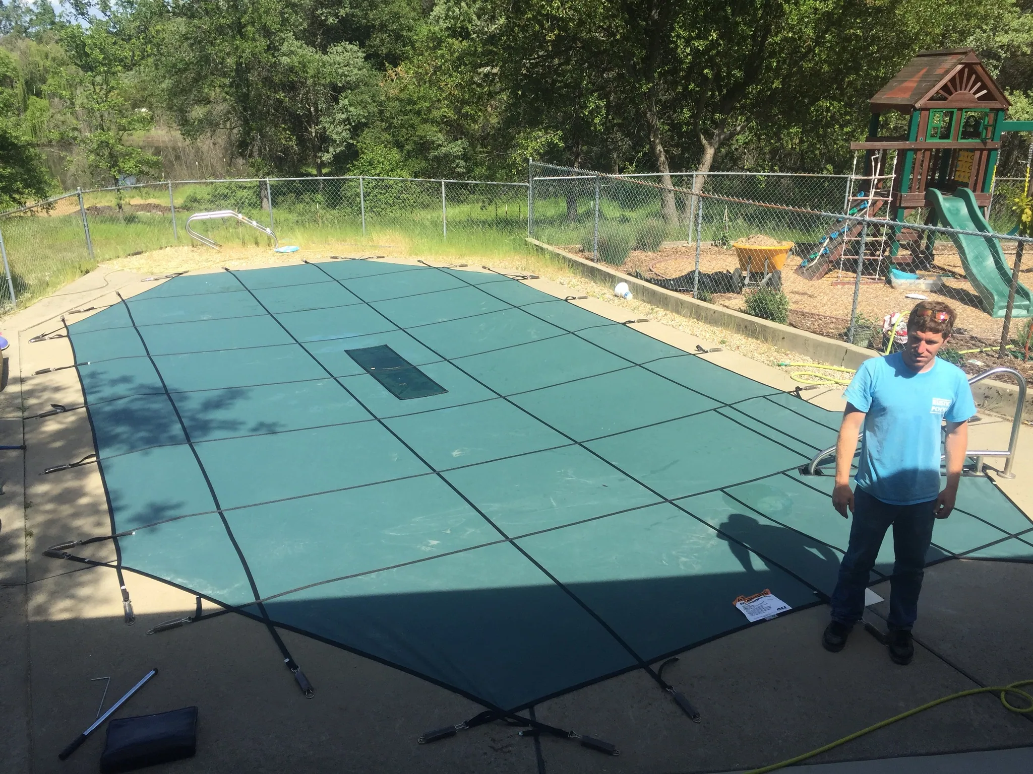 Person standing beside blue mesh pool cover in backyard with playground equipment and chain-link fence in background