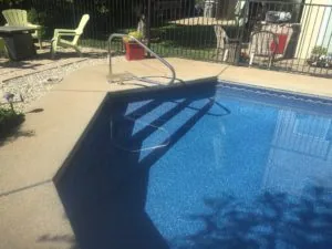 Residential swimming pool with blue water and skimmer hose, concrete deck, metal fence, and patio furniture in background