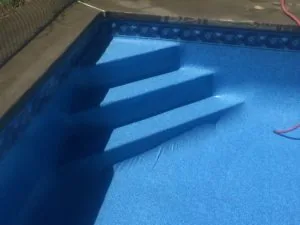 Swimming pool with blue interior surface showing pool steps casting shadows, with concrete deck and red hose visible at edge