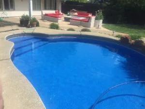 Backyard swimming pool with blue water and concrete deck, red and tan patio furniture in background near house