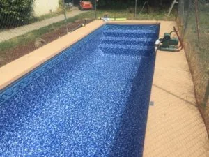 Rectangular residential swimming pool with blue tile interior and tan concrete deck, surrounded by chain-link fencing