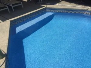 Clear blue swimming pool with decorative tile border and concrete deck, lounge chairs visible in background