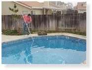 Person using a pole tool to clean a backyard swimming pool with wooden fence in background