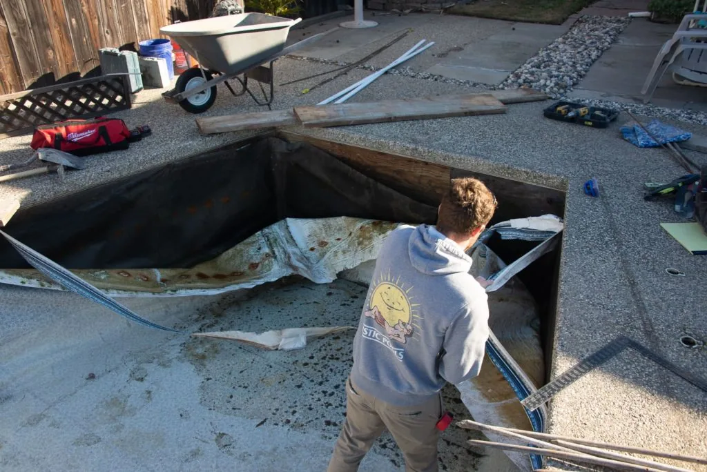 Worker standing in empty in-ground pool during construction or renovation with tools and materials scattered around the concrete surrounding area