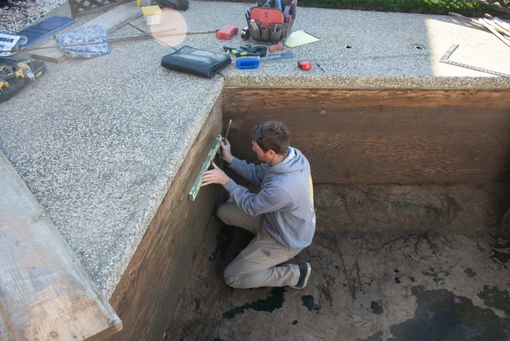 Man crouching inside wooden-framed pool basin, measuring or inspecting interior wall