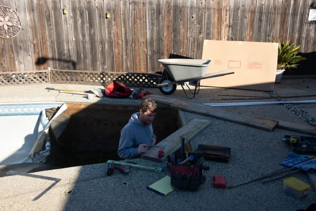Worker in pool pit with tools and materials, wooden fence and wheelbarrow in background