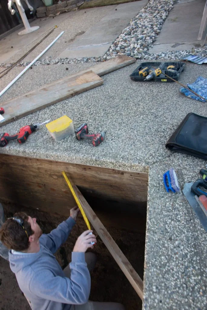 Worker measuring wooden frame opening in gravel surface with construction materials and tools nearby