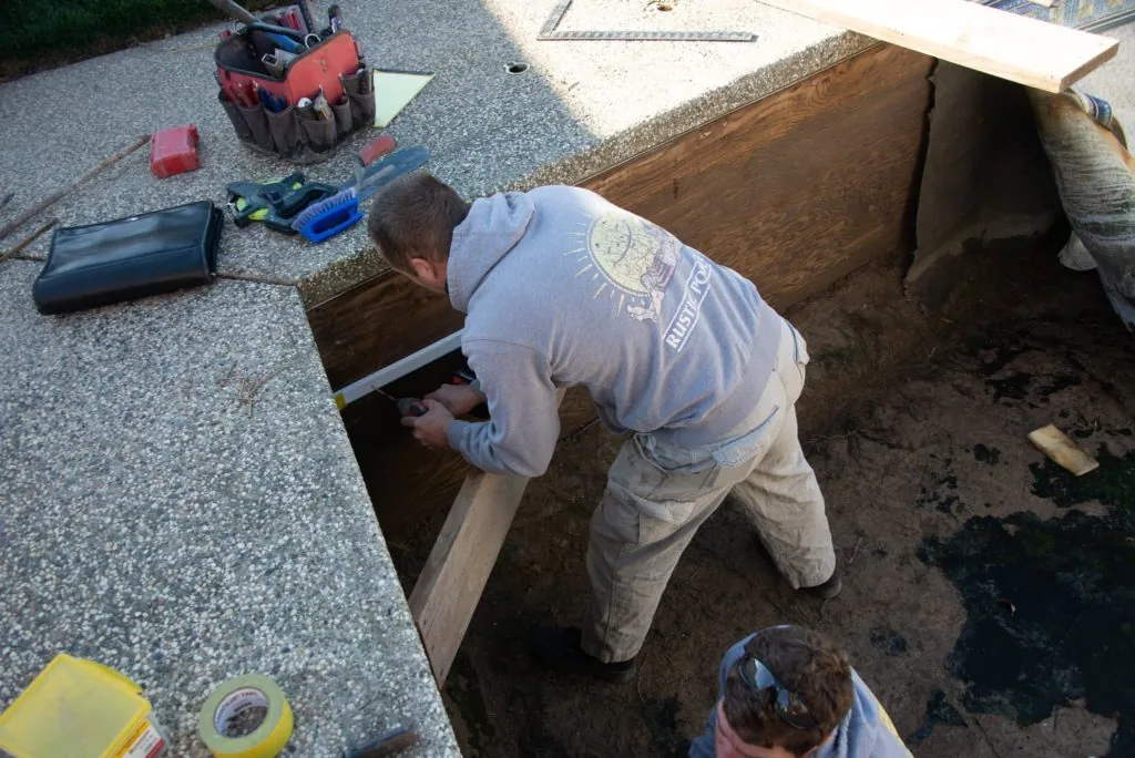 Worker bending into wooden foundation pit during construction or repair work, with tools and equipment on gravel surface nearby