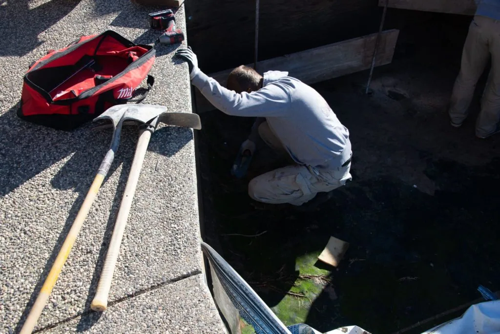 Person in white shirt crouching by pool edge with red net and cleaning tools on concrete deck