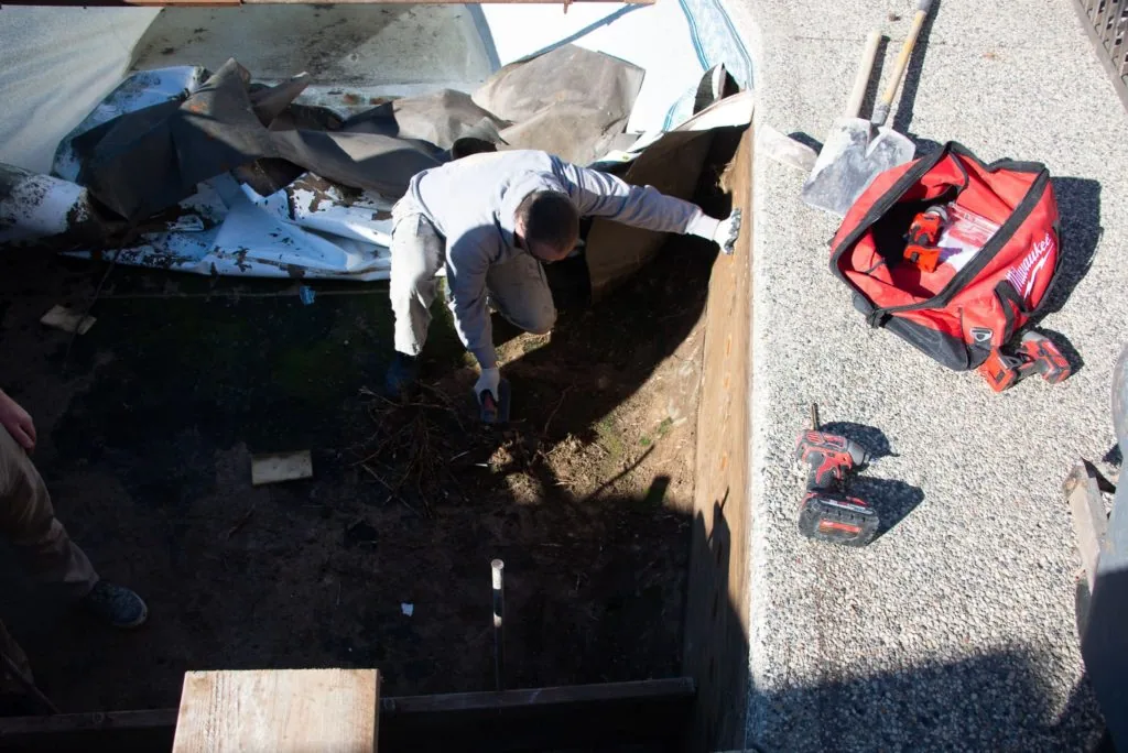 Worker leaning into excavated pool pit with tools and red equipment bag on concrete surface