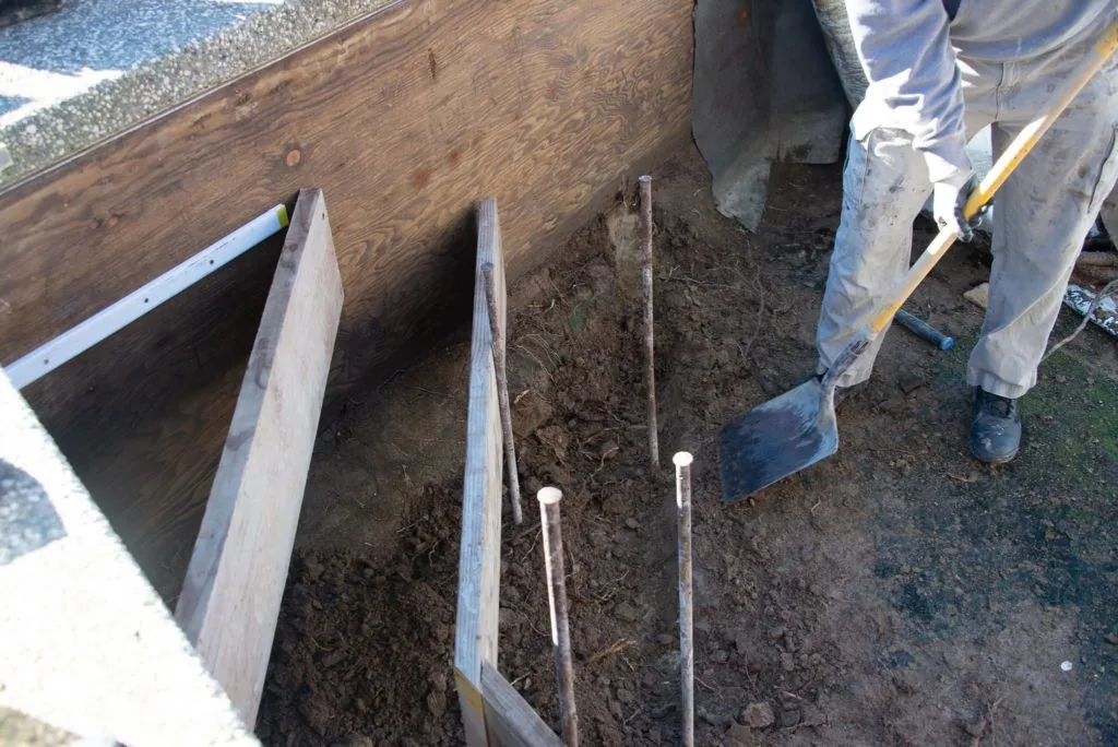 Worker in white pants standing beside wooden framed excavation pit with soil and metal stakes for pool construction