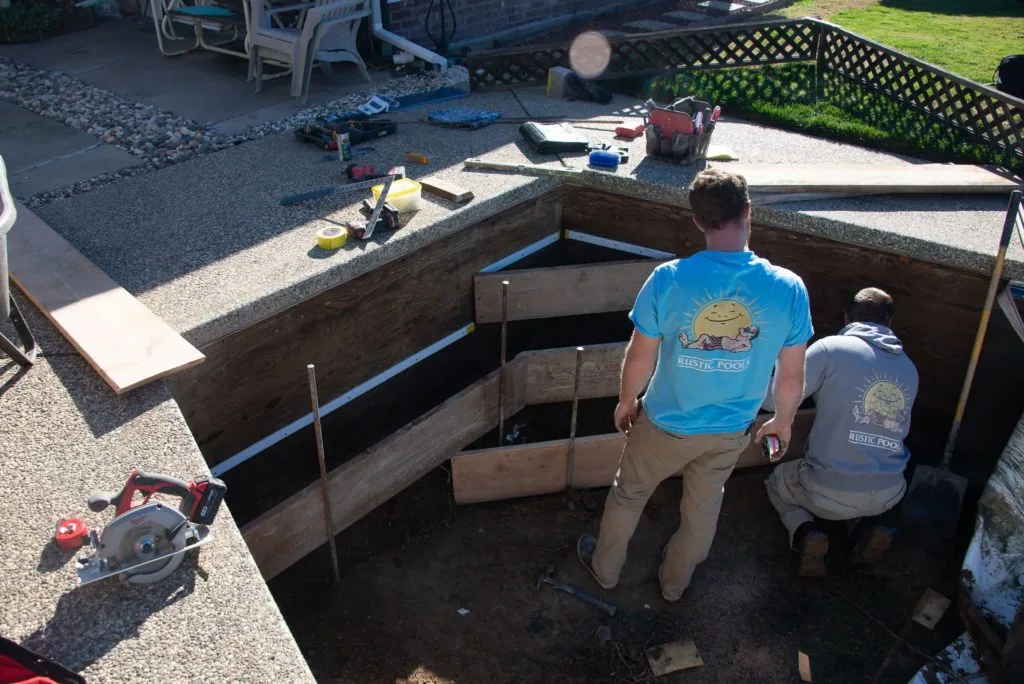 Two workers in branded shirts standing in an excavated pool foundation with concrete walls and wooden framing during construction