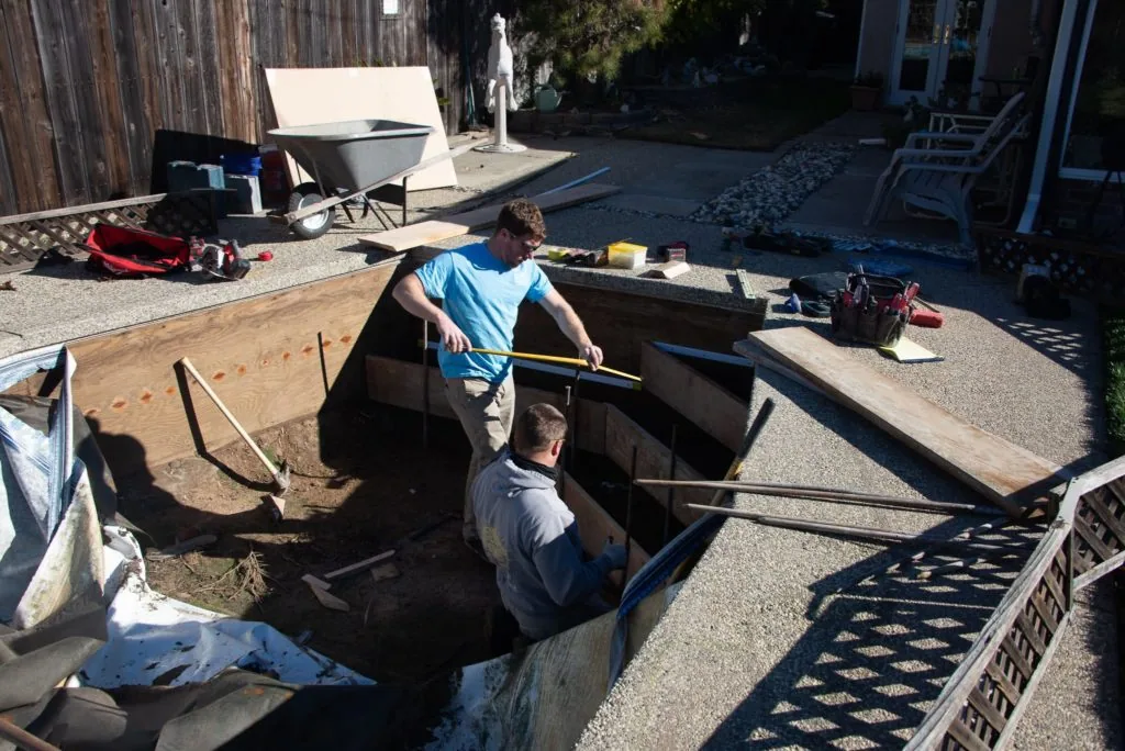 Two workers in a backyard excavation pit with wooden framing, surrounded by tools and construction materials