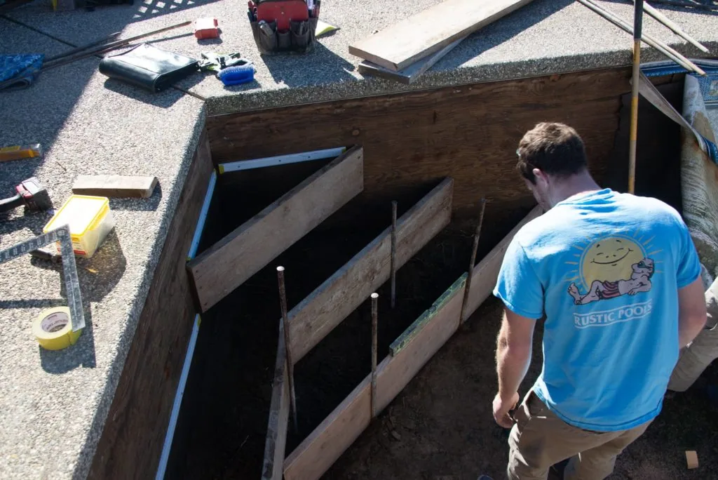 Worker in blue shirt standing at basement egress window opening with wooden frame and concrete steps during construction