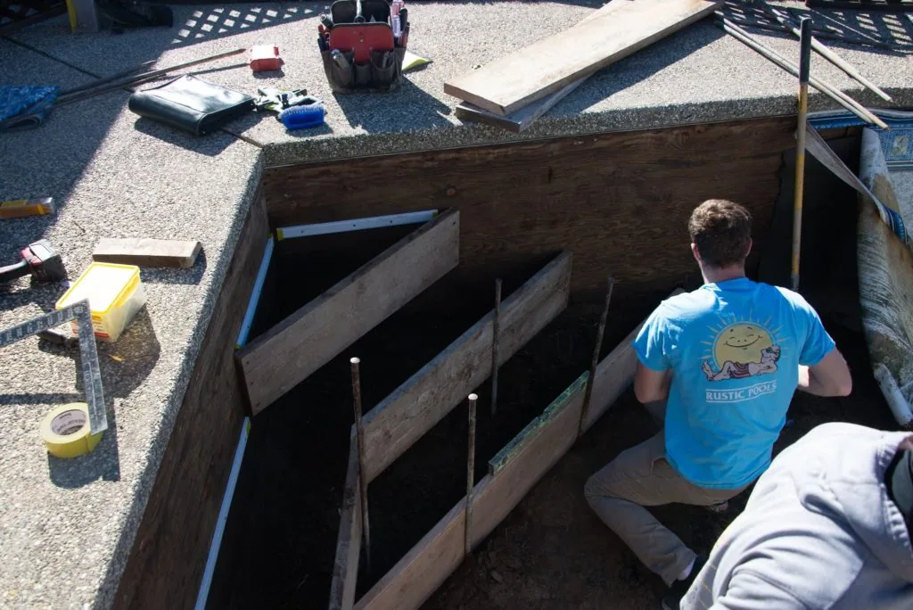 Worker in blue shirt descending into wooden-framed basement excavation with concrete pad and tools visible above
