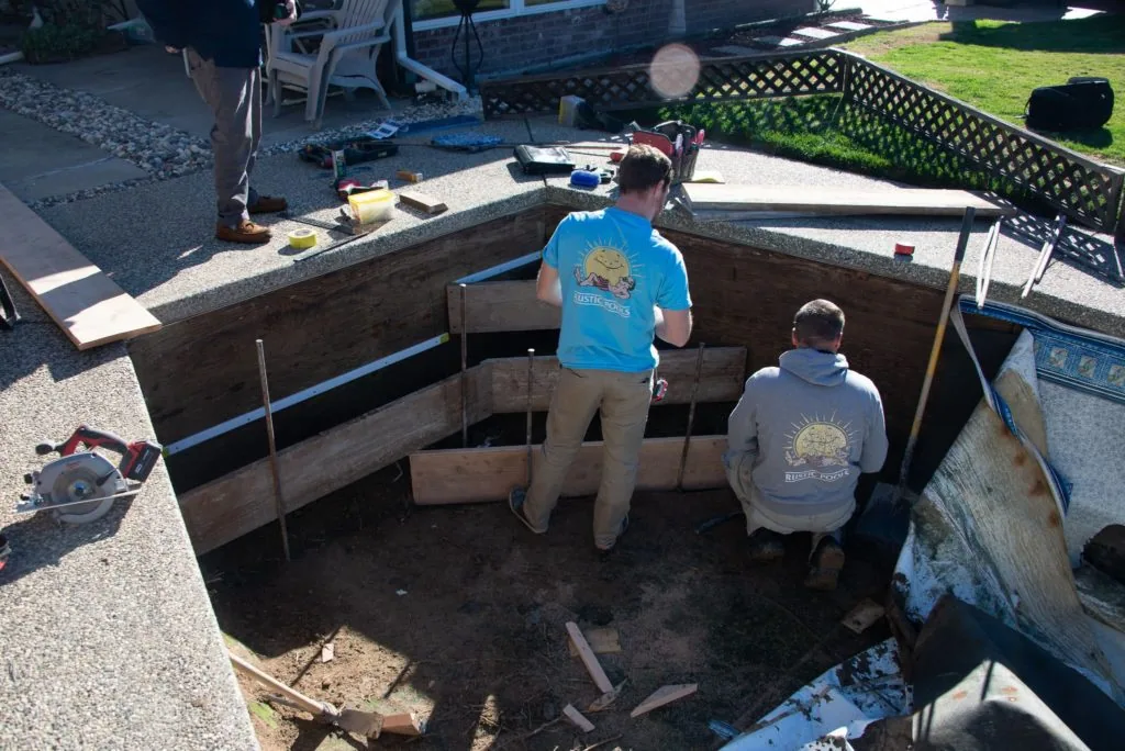 Two workers in branded shirts standing in an excavated pool foundation with wooden framing and tools visible