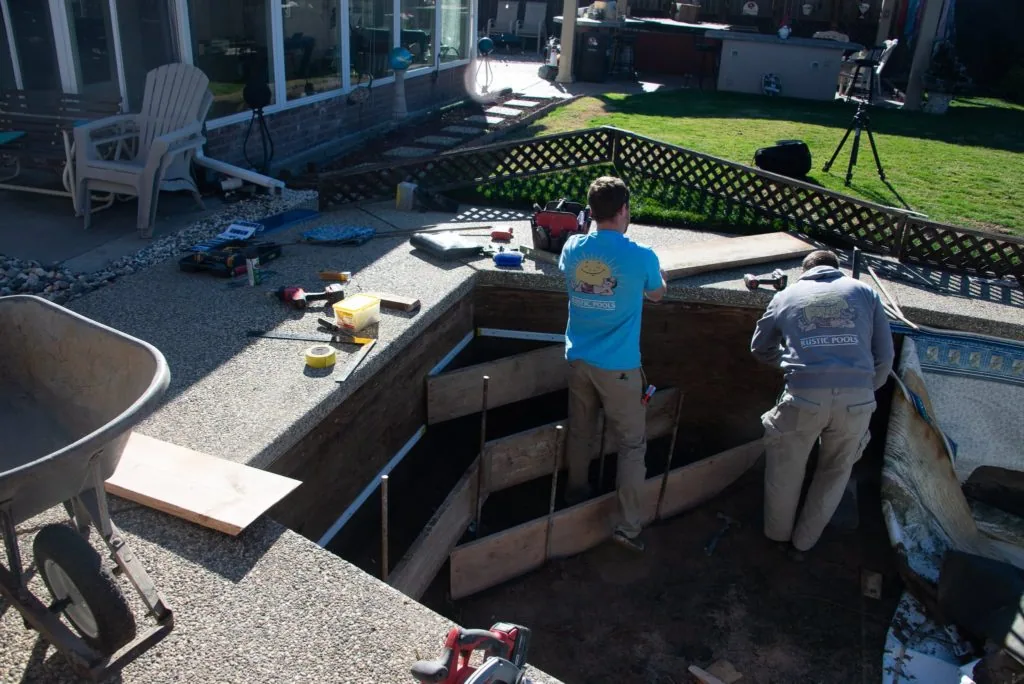 Two workers standing in an in-ground pool under construction with gravel, tools, and equipment visible around the backyard site