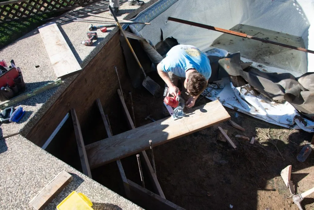Worker in blue shirt crouching in excavated foundation pit with wooden framing and construction materials surrounding the site