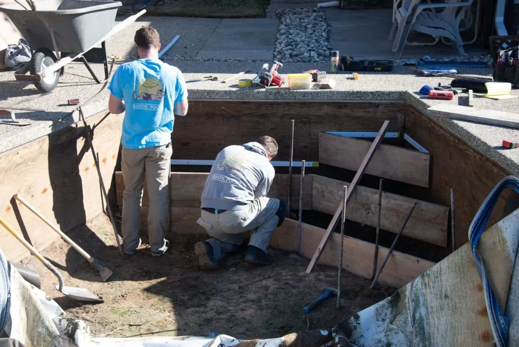 Two workers performing maintenance work inside an empty concrete pool pit with tools and materials visible around the area