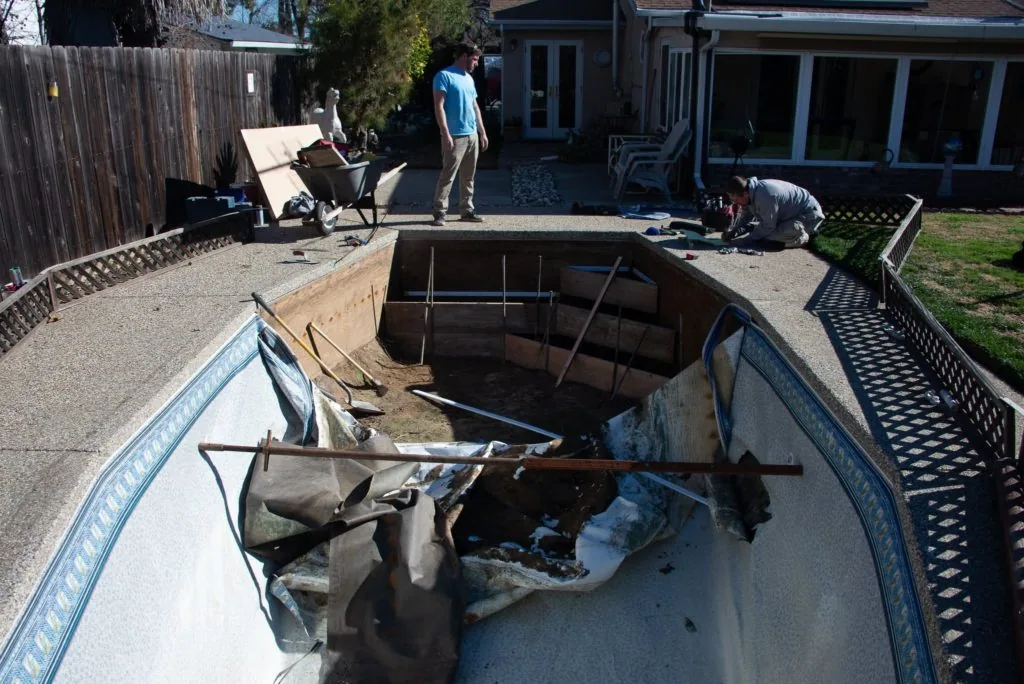 Drained backyard swimming pool during renovation with workers and equipment visible