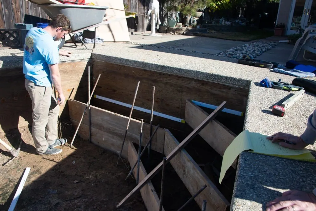 Worker in blue shirt standing beside concrete pool construction with wooden interior framing and metal supports