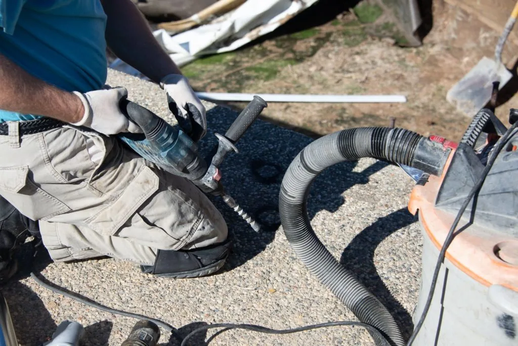 Person in work gloves holding black corrugated pool hose on gravel ground near equipment