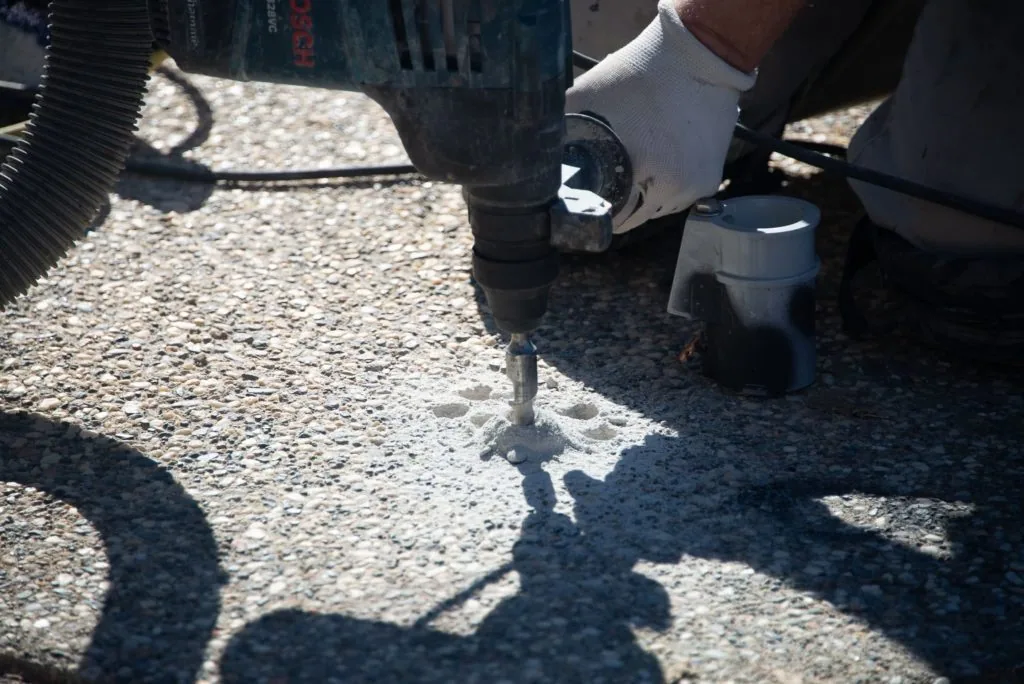 Person in white glove operating pneumatic air hammer drill on gravel surface near construction equipment