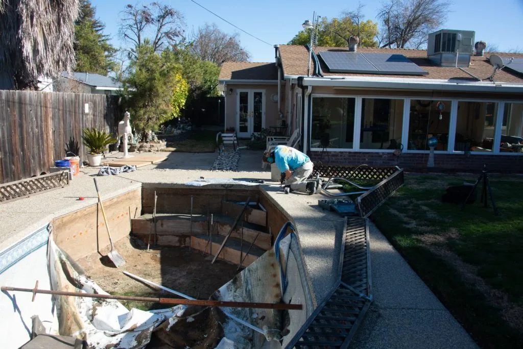 Residential backyard with in-ground pool under construction, worker bending over excavation, house with large windows and solar panels visible