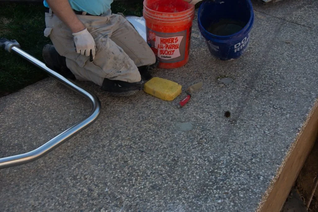 Worker in protective gloves scrubbing concrete surface with yellow sponge near paint buckets and hose during pool maintenance