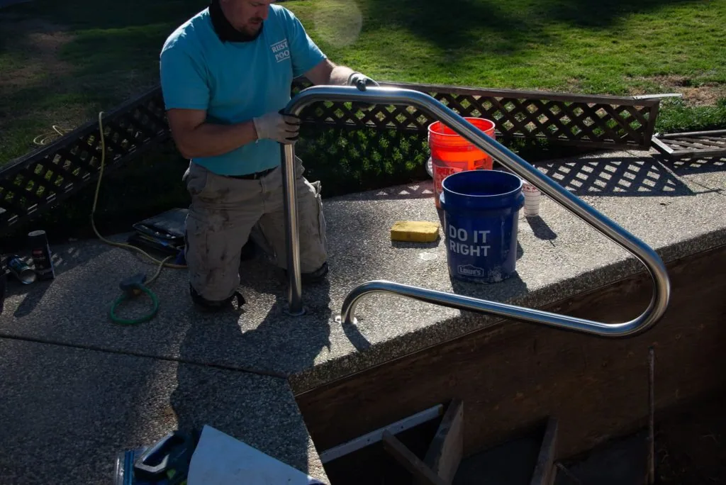 Man in blue shirt installing metal railing on concrete pool deck next to bucket and tools