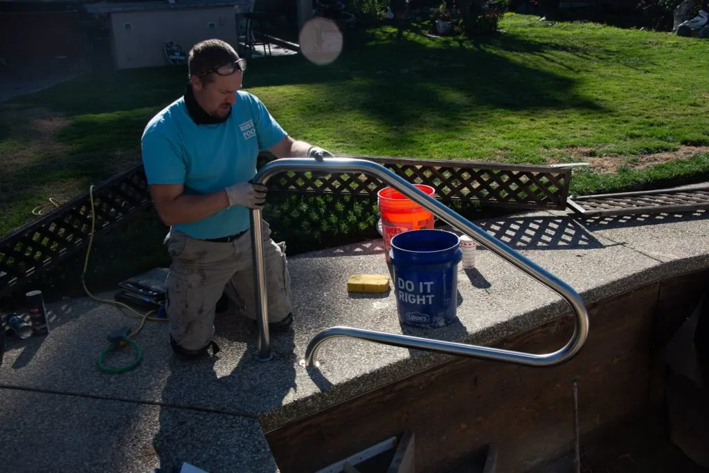 Man in blue shirt working on pool equipment with metal tubing and blue bucket on concrete