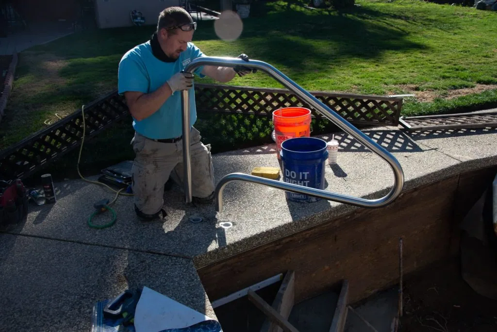 Man in blue shirt installing metal railing on pool deck with paint buckets nearby