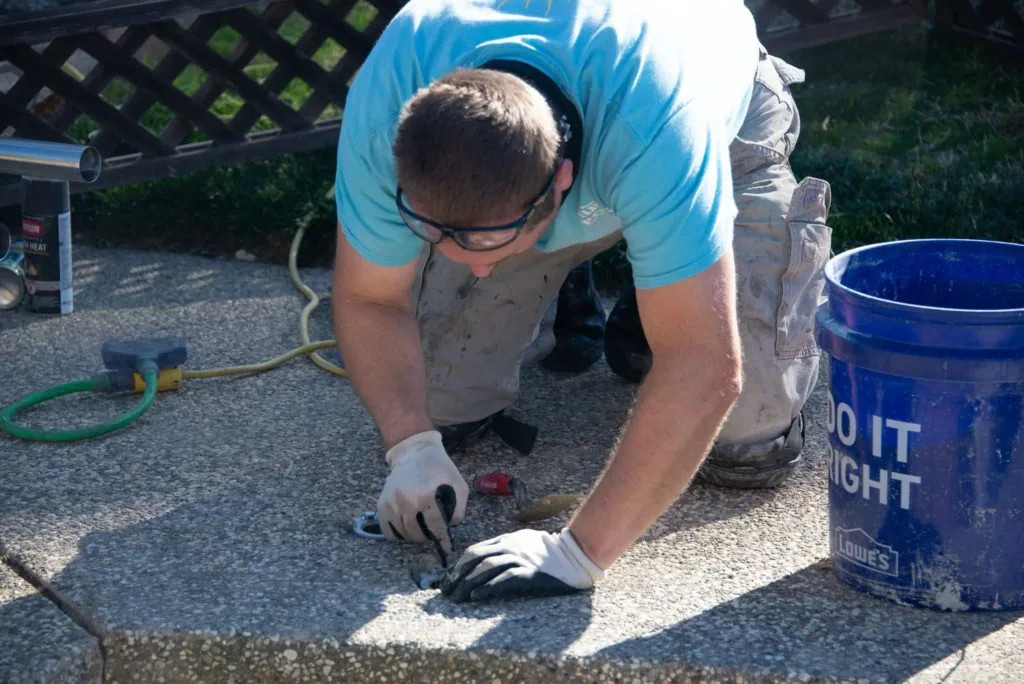 Man in blue shirt and glasses working on concrete surface with trowel, blue bucket nearby