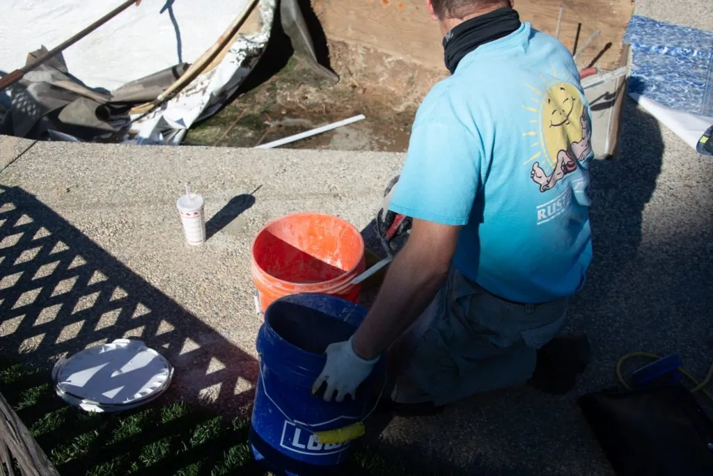 Person in blue shirt crouching beside orange and red buckets on concrete surface with pool equipment visible