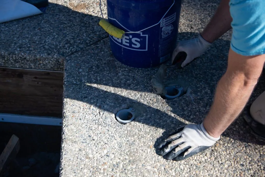 Person in white glove holding blue bucket with yellow object near gravel surface with drilled holes