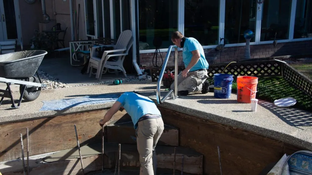 Two workers performing maintenance or construction work on a backyard pool, with tools and equipment visible nearby
