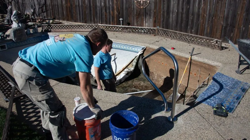 Worker in blue shirt and tan pants leaning over an in-ground pool under construction in a backyard with wooden fence