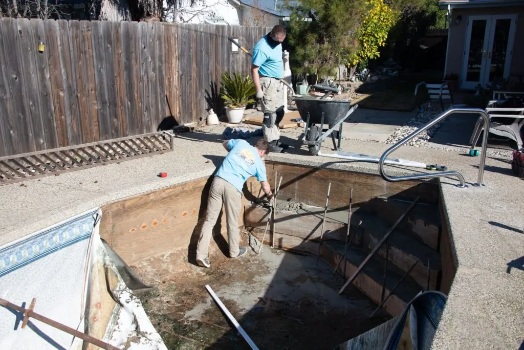 Two workers preparing a backyard pool renovation with equipment and materials visible on a sunny day